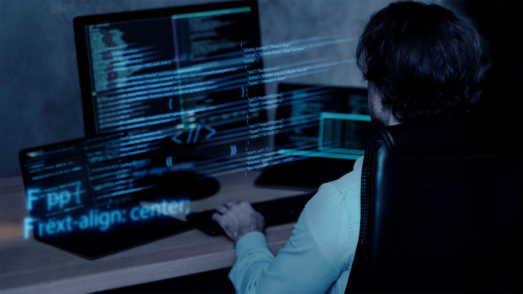 Mulher branca sentada de costas e de frente para um computador com a tela repleta de códigos.